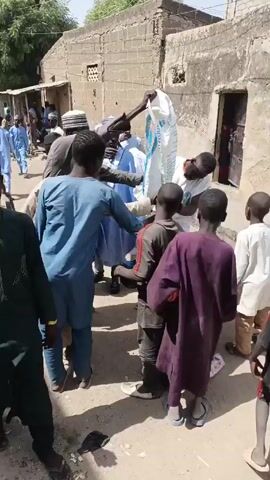 The video depicts a scene in what appears to be a village or town, with buildings constructed of brick or concrete visible in the background. A group of people, predominantly men and boys, are gathered around a man who is holding a large white sack with blue writing. The man appears to be distributing the contents of the sack, as he pours some into the hands or containers of those gathered around him. The men are wearing a mix of traditional clothing and casual wear, and some of the boys are carrying containers or bags. The atmosphere appears to be orderly.