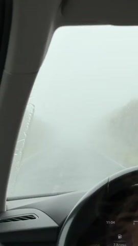 The view from inside a car shows a road obscured by heavy rain and fog. A single windshield wiper blade sweeps across the windshield. The car's dashboard is visible, displaying a time of 11:04. The driver's hands are not visible, only the steering wheel. The car appears to be in motion.