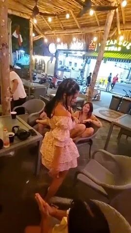 Here's a description of the video:

The video shows a young woman dancing in what appears to be an outdoor seating area of a restaurant or cafe. She is wearing a pink floral two-piece outfit and has dark hair. Several other people are around her, including a woman lounging in a chair behind her and a person holding a phone taking a video. The camera focuses on the dancing woman, who is smiling and interacting with the camera. The environment is well-lit with string lights and other outdoor lighting. There are tables and chairs visible, and in the background, there are glimpses of a street with shops and pedestrian activity.