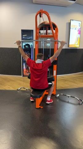 The video shows a man in a red t-shirt and black shorts performing a lat pulldown exercise on an orange weight machine in a gym. He is seated facing the machine, gripping a bar with his hands extended above his head. His back is to the camera, and he is leaning back slightly as he pulls the bar down towards his chest. The gym environment is visible in the background, with other exercise equipment and a promotional poster on the wall. The floor is dark with an orange accent.