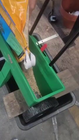 A person pours grass seed from a yellow bag into a green lawn spreader. The spreader has black handles and a black wheel at the bottom. It sits on top of a black tray, which is on a wooden plank. The floor appears to be concrete.