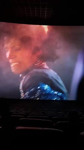 The video shows a movie screen inside a cinema. The film appears to be a biopic about Michael Jackson. The film depicts a pyrotechnic malfunction, or maybe a fictionalized event where Jackson is set on fire during a performance. The seats in the cinema are empty except for one occupied seat visible in the lower right. The viewer is sitting in the cinema, recording the screen. The shot cuts to reporters at a press conference. Then, the shot cuts back to the depiction of the onstage pyrotechnic malfunction.
