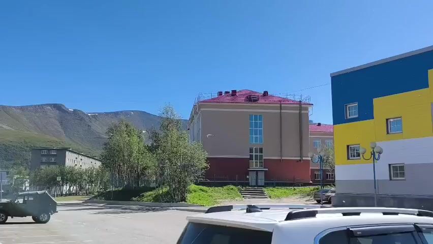 This is a horizontal, eye-level shot of a small town on a clear day. A bright blue sky dominates the upper half of the frame, with hazy, muted green mountains in the background on the left. In the mid-ground, a large, tan building with a pink roof and a blue-accented window section is visible. To the right of this building is a multi-story structure with bold blue and yellow sections. In the foreground, the top of a white SUV is visible. A dark green off-road vehicle is parked to the left of the tan building, and another black SUV is parked behind it. A few trees with green foliage are scattered throughout the scene, with some green grass and patchy dirt visible between the buildings and vehicles. The overall impression is one of a remote, possibly northern, location with a mix of older and more modern architecture.