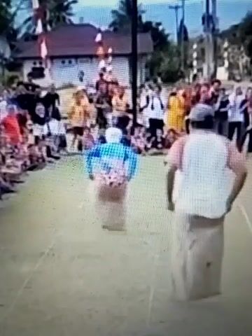 The video shows an outdoor sack race with several participants hopping in burlap sacks. A crowd of spectators, including children and adults, are gathered on both sides of the race course, some sitting and some standing. In the background, there are buildings and trees, and a tall pole with red and white flags is visible. The ground is covered in grass, and white lines mark the boundaries of the race. The overall atmosphere appears to be festive and celebratory.