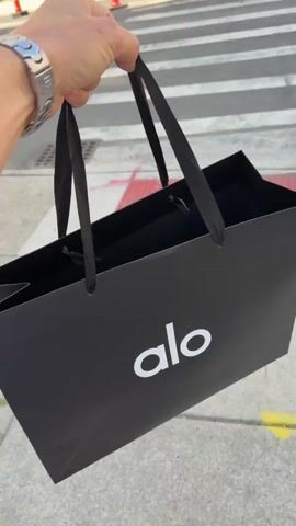 A hand holds a black shopping bag with a white "alo" logo. The bag is carried over a crosswalk with white and grey stripes. The person is wearing a blue bracelet on their left wrist and white socks and athletic shoes. The video then shows a street with buildings on either side and traffic cones on the right side of the road.