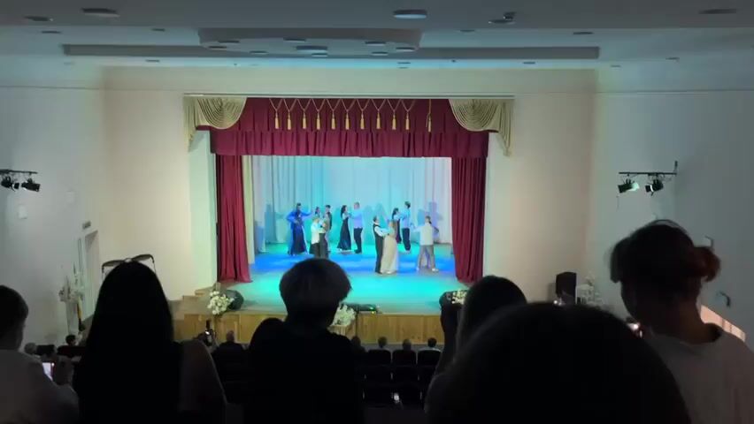 This is a video recording of a dance performance taking place on a stage in what appears to be an auditorium or community hall. The stage is decorated with a maroon curtain and lighter-colored drapes, with decorative gold tassels along the top. Several people, dressed in formal attire, are dancing in pairs. The lighting on the stage shifts in color, cycling through blues, greens, and purples, creating a dynamic atmosphere for the performance.

In the foreground, several audience members are visible from behind, obscuring their faces. They are seated in what seem to be rows of chairs, and some are holding up phones, suggesting they are recording the event. The overall mood appears to be one of polite appreciation for the performance. The setting suggests an indoor event, likely in the evening due to the artificial lighting. There is no visible text in the video. The location context is Pishchane, Ukraine, which implies the performance is a local cultural event. The participants' clothing suggests a formal or celebratory occasion, possibly a school event or a local dance competition.