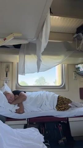 A person is sleeping on a bunk bed in a train compartment. The person is covered by white sheets and wearing patterned shorts and white socks. The train compartment has a window with a view of trees and a bright sky. There are shelves above the bunk bed with items stored on them, and bags are visible underneath the bunk bed.
