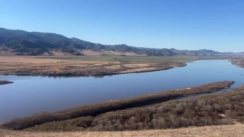 The video shows a wide river flowing through a valley with mountains in the background under a clear blue sky. The river has several islands covered in trees and shrubs. The valley is mostly dry grassland with some patches of green fields. The foreground shows a grassy hillside. There are no people visible in the scene.