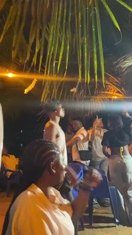 The video shows a group of people dancing and socializing at an outdoor party at night. They are gathered around a DJ booth, with palm trees overhead. Most people are standing, but one woman sits in the foreground. People are wearing casual clothes and dancing energetically. The video is dimly lit with a warm light.