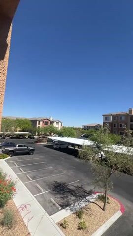 The video shows a bright, sunny day with a clear blue sky. The camera is positioned at an angle looking down on an apartment complex parking lot. In the foreground, there are parking spaces marked with white lines on asphalt. A small, bare-branched tree with a rocky bed sits in the corner of the parking lot, bordered by a red curb. To the left, a black truck is parked. Further back, a long, covered carport with a white roof stretches across the parking lot, sheltering several vehicles. Beyond the carport are residential buildings with multiple stories, featuring tan and brown exteriors, balconies, and windows. Green trees and landscaping are visible between the buildings. In the distance, more residential buildings and trees are visible under the bright sky. The sun is high in the sky, casting shadows.