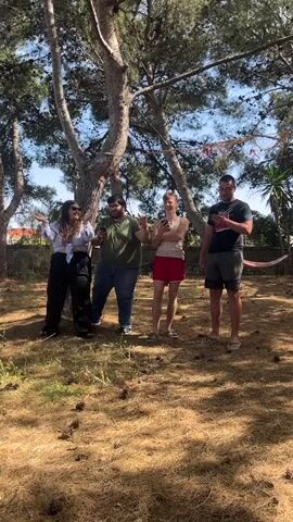 Four individuals, two women and two men, stand in an open, grassy area beneath trees. The ground is covered in dry pine needles and cones. They all hold phones and are looking at them, then they begin clapping. The women are on the left, one wearing sunglasses and a white shirt, the other in black and a dark green shirt. The men are wearing short-sleeved shirts and shorts. A hammock is visible in the background. The sky is clear and blue.