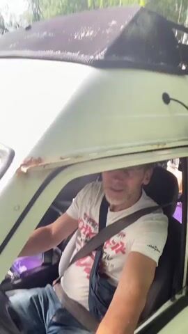 A man wearing glasses, a white t-shirt with a graphic print, and blue denim overalls is driving a vehicle. He is looking forward and smiling. Another person is visible in the passenger seat. The vehicle appears to be an older model van or camper with a roof rack. The scene takes place outdoors, with trees visible in the background. The video is shaky, suggesting movement.