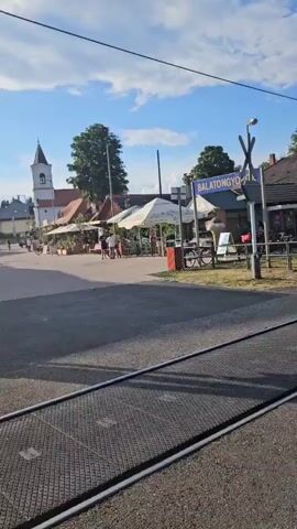 The video begins with a sign that reads "BALATONGYÖRÖK." A railroad crossing with lights and arms is visible in the foreground, with tracks extending into the distance. Several bicycles are parked near the crossing. In the background, there are buildings with awnings, suggesting shops or cafes. People are seen walking and riding bicycles in the street. A white church steeple rises above the buildings on the left side of the frame. The sky is blue with scattered clouds, and the sun appears to be shining, indicating daytime. The overall scene depicts a street in a town, likely a tourist area, with a mix of infrastructure, businesses, and pedestrian activity.