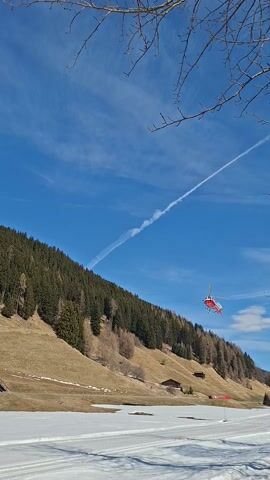 The video captures a red and white helicopter flying across a mountainous landscape under a clear blue sky.

At the beginning of the video (0:00), the helicopter is seen in the upper mid-left of the frame, flying from right to left. Below it is a dense, dark green forested hillside, while to the right, a wide, snow-covered slope is visible. The sky is a vibrant blue, with a faint white contrail stretching diagonally across it from the upper left towards the lower right. Some bare tree branches are visible in the extreme upper left corner of the frame.

As the video progresses (0:01 - 0:02), the helicopter continues its trajectory, moving further left and slightly descending. The contrail remains a prominent feature in the sky.

By 0:02, the helicopter has moved lower in the frame and is now over the brown, partially snow-dusted hillside, which is sparsely covered with trees. A multi-story building with a brown facade and a dark roof becomes visible on the hillside to the right of the helicopter. The snowy slope on the far right continues to be a consistent element.

In the final moments of the video (0:03 - 0:04), the helicopter is closer to the bottom of the frame, continuing its flight path to the left and seemingly moving away from the camera. The building is more clearly discernible, nestled on the hillside, and the blue sky with a few scattered white clouds fills the upper left portion of the frame. The distinct sound of the helicopter's rotors is audible throughout the video. The overall impression is a sunny day in a mountain region, possibly a ski resort.