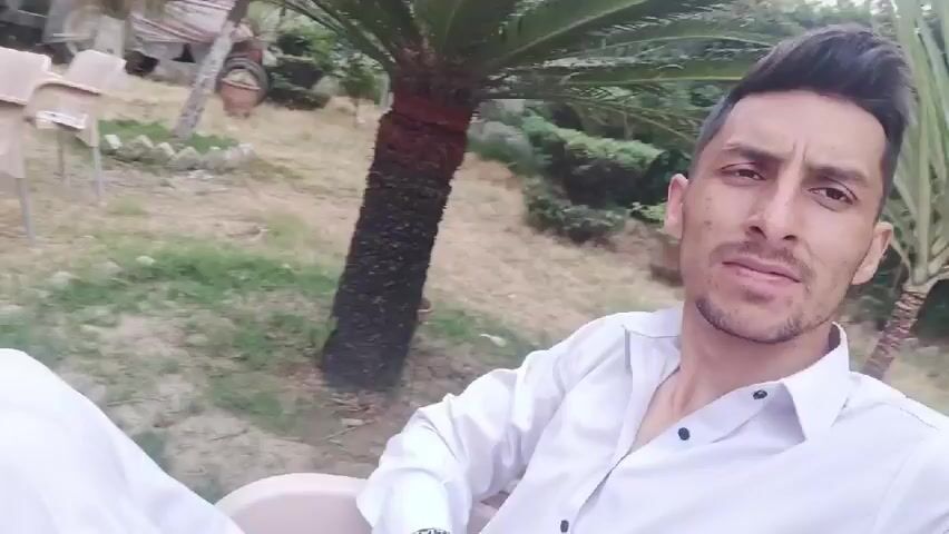 A man wearing a white collared shirt and white pants is sitting outside in a chair, surrounded by foliage. He is looking into the camera with a slight smirk. Behind him are several light-colored plastic chairs, a small palm tree, and a flatbed trailer. The camera pans upward to show a grey cinderblock wall, a door, a variety of shrubbery, and a tall thin tree. The sky is overcast.