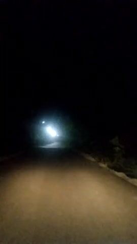 The video captures a nighttime scene from the perspective of a vehicle driving on a dark, unlit road. The headlights of the vehicle illuminate the asphalt surface, creating a pool of warm light that gradually fades into the darkness ahead. In the distance, the bright lights of another vehicle are visible, suggesting oncoming traffic or a stationary source of illumination. The surrounding environment is obscured by the darkness, hinting at a rural or undeveloped area. The overall atmosphere is one of quiet solitude and the focused experience of navigating through the night. The time of day is clearly nighttime due to the complete absence of natural light and the use of headlights. There are no discernible weather conditions, though the darkness could imply a clear or overcast sky. The location is suggested to be Alavari, Georgia, but no specific visual cues confirm this beyond the provided context. There is no visible text in the media.