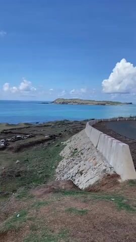 The video shows a coastal scene under a bright blue sky dotted with white clouds. In the distance, a small, rocky island sits in the turquoise water. The foreground consists of a grassy area with stepped terraces, leading down to a rocky shoreline.  

A concrete barrier runs alongside a road. As the camera pans, people can be seen walking and taking pictures near the water's edge.  

The camera continues to pan, revealing a small area set up with chairs and a table overlooking the ocean.  A few people are seen standing on the cliff's edge.