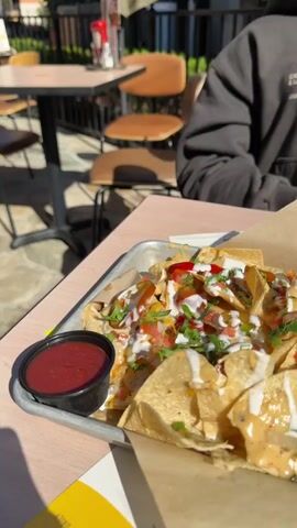 The video shows a close-up of a tray of nachos on a table, likely at an outdoor restaurant. The nachos are covered in cheese, salsa, white sauce, and chopped cilantro. There is a small container of red sauce on the side. A person wearing a grey sweatshirt sits in the background. The table and chairs of the restaurant are visible but out of focus. The lighting suggests it is daytime and sunny.