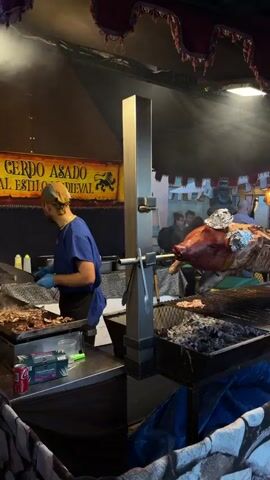The video shows a medieval fair with a whole roasted pig on a spit being cooked over hot coals. A man in a blue shirt is cutting up other cooked meat at a counter. A man in a white shirt and a red and white apron carries a rolled-up umbrella across a paved area. People walk along the street lined with fair stalls decorated with red and white pennants. Buildings are visible in the background.