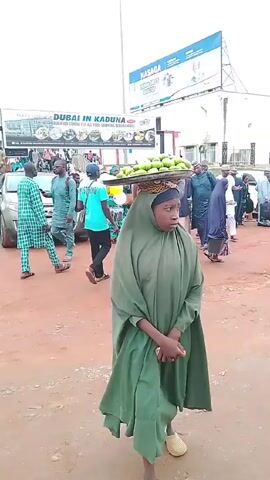 The video depicts a busy street scene in what appears to be a town in Nigeria. A young woman in a green hijab is walking toward the camera carrying a tray of green fruit, likely mangoes, balanced on her head. She appears to be selling the fruit. Around her, many people are walking in various directions, some wearing traditional clothing, others in more casual attire. There are parked cars along the side of the street. A large billboard overhead advertises "Dubai in Kaduna," suggesting a shopping center or event. The environment looks dusty and urban, with a red dirt or gravel surface underfoot.