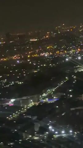This is a vertical, shaky video taken at night from a high vantage point, possibly an airplane, looking down at a sprawling city. Numerous lights are visible, suggesting a densely populated urban area. The lights are a mix of white, yellow, and some colored lights, possibly from streetlights, buildings, and vehicles. The landscape appears to be hilly or mountainous, with the city lights spread across the slopes and valleys. The overall impression is of a vast, illuminated cityscape at night, shrouded in darkness. There are no visible people or distinct objects, and no discernible actions or interactions taking place. The environment is dark with the city lights providing the only illumination.
