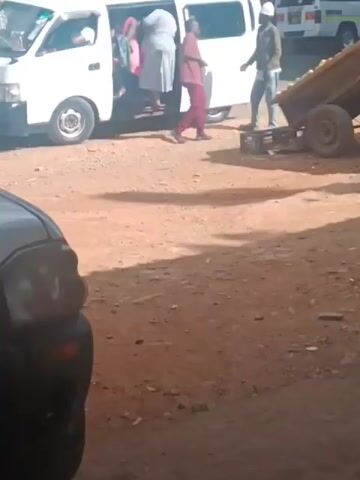 The video shows a somewhat chaotic scene in a dusty, possibly public, area.  A white minivan is parked, and several people are either getting in or out of it. One person wearing a white garment and another carrying a pink bag are visible in the doorway of the van. To the right of the van, two men are standing near a two-wheeled cart filled with what appears to be produce. One man is wearing a white hard hat.  A person in red clothing is walking away from the van.  A black car is parked in the foreground on the left, partially obscuring the view.  The ground is dirt and gravel. Other vehicles can be seen in the background.