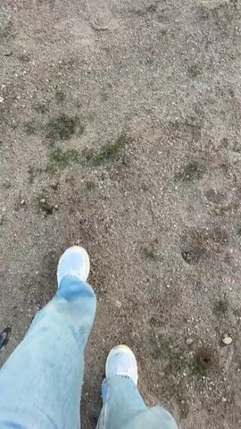 The video opens with a first-person view of someone walking on a dirt path, showing their light blue jeans and a light blue and white striped shoe. A metallic clip hangs near their left leg. As the camera pans up, a breathtaking scene unfolds: dozens of colorful hot air balloons fill the sky and are spread across a dirt ground in Göreme, Turkey, at sunrise.

Many people are gathered on the ground, observing the balloons. Some balloons are already airborne, gracefully ascending into the dawn sky, which transitions from soft blue overhead to vibrant oranges and yellows near the horizon. Others are still on the ground, either partially inflated or fully upright, preparing for takeoff.

A notable object on the ground is a vintage pink convertible car, positioned near some of the grounded balloons. Visible text on the balloons includes "URGUP" on a large orange balloon, "KAYA HOTEL" on a white balloon, and "MEGA" on another, possibly "MEGA GÖREME". The scene captures the iconic and serene hot air balloon spectacle during a beautiful sunrise in Cappadocia.