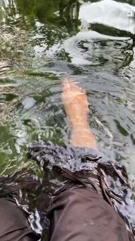 The video shows a first-person perspective of a person's legs and feet submerged in shallow, clear water, likely a stream or creek. The water has a greenish tint due to algae or submerged vegetation, and sunlight reflects off the rippling surface. The person is wearing dark, waterproof pants that are wet. The individual is moving their feet and legs, causing ripples and disturbances in the water. The surrounding environment appears to be natural, with hints of green foliage visible through the water's surface in the background. The overall impression is one of a person enjoying a moment of cool relief in a natural water setting.