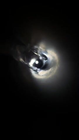 The video shows a close-up, blurry, and mostly dark view of a liquid being poured into a container. Light reflects off the liquid's surface, creating a bright, almost blinding glare, which is surrounded by darker, swirling shades. The environment is obscured by darkness. The pouring action is central, suggesting the focus is on the liquid and its movement.