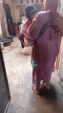 A child is being carried on the back of an adult. The adult is wearing a light purple robe and a hijab. The child is wearing a red shirt and a patterned wrap. The adult is walking on a concrete path, with a wall on the left and a building in the background. Another person is bent over, handling a white bag. A motorcycle is visible in the background. The scene appears to be an outdoor or semi-outdoor area, possibly a yard or alley.