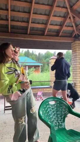 The video opens with a man in a dark jacket and shorts walking away from the camera outdoors under a covered patio. A child in a dark hooded jacket is playing on a red toy. To the left, a person in a light-colored top is partially visible. Further in the background, two women are seated at a table, and another person is standing. The environment appears to be a backyard with houses and trees in the distance. As the video progresses, the camera pans to the right, revealing a sunset with clouds. Another person, wearing a white vest over a pink top and white pants, is facing away from the camera. Smoke is visible in the foreground, suggesting a hookah or similar device is being used. The scene depicts a casual outdoor gathering.