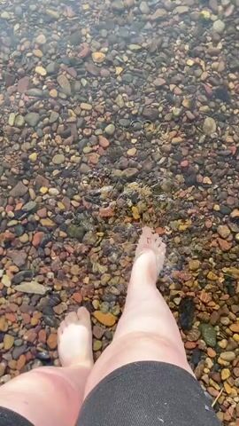 A first-person perspective video shows feet and legs wading into a shallow body of water with a rocky bottom. The water is clear and reflects sunlight, revealing the colorful pebbles beneath the surface. The person's feet are bare and appear to be pale. The person is wearing dark shorts. The water is rippling around the legs and feet as the person moves forward.