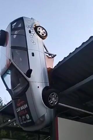 The video shows a silver car suspended upside down from a metal structure, possibly a roof or awning. The car has red and white stripes on its side and a large black sticker on the rear window that reads "CAR WASH UNTILA" with icons for price, time, and quality. The background is a bright sky and some trees are visible below. The car appears to be a promotional display for the car wash.