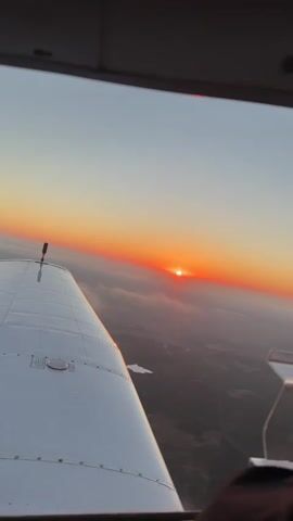 This is a first-person perspective video taken from inside an aircraft. The camera looks out a window towards the horizon, where a vibrant orange sunset is visible. The sun is a bright, glowing disc just above a layer of scattered clouds. Below the clouds, the landscape is indistinct but appears to be rural, with some patches of darker vegetation and lighter open areas. A portion of the aircraft's wing, which is white and metallic, occupies the left side of the frame, extending towards the foreground. A small antenna or sensor protrudes from the wing. The sky transitions from a pale blue at the top to a deep orange and red near the horizon. The overall mood is serene and awe-inspiring, capturing the beauty of a sunset from an elevated viewpoint.