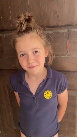 The video features a young girl with her hair in a messy bun, wearing a dark blue polo shirt with a yellow smiley face patch on the left side of the chest. She is standing in front of a wooden door. The girl appears to be speaking and smiling, with a cheerful and animated expression. Her movements are playful, as she shifts her weight and gestures with her hands. The lighting suggests it is daytime, likely outdoors due to the natural light illuminating her face. The wooden door behind her has visible grain and appears to be a rustic or aged design, possibly from a home or an older building. The setting is likely Castelfranco di Sotto, Italy, based on the provided context. No discernible text other than the smiley face on her shirt is visible.