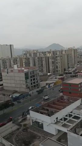 This media file shows an aerial view of an urban landscape with a large plume of black smoke rising into the sky. The setting appears to be Breña, Peru, as indicated by the location context.

**Subjects:**
*   **Scene/Setting:** The scene is an urban environment with numerous tall residential buildings, industrial-looking structures with large roofs, and a road with traffic. In the background, hills or mountains are visible under an overcast sky.
*   **Objects:** Buildings of various heights and architectural styles are prominent, including modern apartment complexes and what appear to be warehouses or factories. Vehicles, including cars and trucks, are visible on the road. The most striking object is the immense column of black smoke.
*   **People:** A few people can be seen on the street, appearing to be observing the smoke plume.

**Activity or Event:**
The primary event depicted is a large fire, evidenced by the substantial amount of black smoke billowing upwards. The smoke is so dense and extensive that it dominates the upper portion of the frame, obscuring the sky and suggesting a significant blaze. The presence of people on the street might indicate they are reacting to the fire, possibly evacuating or watching the emergency response.

**Notable Details:**
*   **Time of Day/Weather:** The sky is overcast and appears to be daytime, but the light is diffused, suggesting cloudy or possibly late afternoon conditions. The weather seems to be neutral or slightly somber due to the cloud cover and the dramatic smoke.
*   **Location Cues:** The combination of dense residential high-rises and industrial structures, along with the mountainous backdrop, is consistent with urban areas in Peru, and specifically Breña.
*   **Emotions:** The visual of thick, black smoke rising from a city typically evokes feelings of concern, alarm, and urgency. The scene suggests a potentially dangerous situation.
*   **Visible Text:** There is no discernible text visible in the media file.