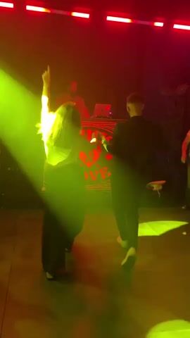 The scene is a dimly lit nightclub with a DJ standing behind a patterned console on an elevated platform. The lighting is primarily red, with periodic green spotlights sweeping across the dance floor. A couple is dancing in front of the console, their movements and faces obscured by the dark lighting. Other patrons are visible in the periphery, though their features are similarly indistinct. The floor is composed of large tiles.