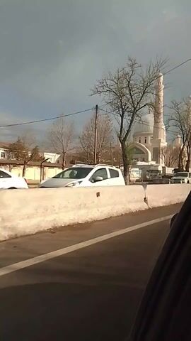 The video, taken from within a moving vehicle, showcases a large, ornate mosque with a dome and two minarets. The mosque is painted a light cream color and stands behind a low, gray concrete barrier along a road. Several trucks are parked along the side of the road in front of the mosque, and other cars can be seen in the background. Bare trees frame the mosque on both sides, and a building with a sign written in Cyrillic script is visible to the left. The sky is overcast, and the overall impression is one of a cold, urban environment.