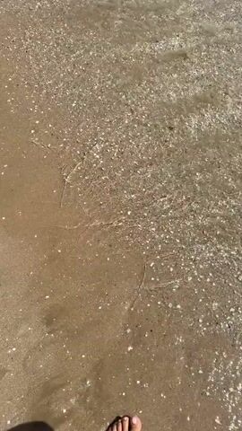 A first-person perspective video shows feet walking on a sandy beach as small waves approach and recede. The water is clear and the sand is covered in small shells. In the distance, a city with many buildings is visible along the coast. Several people are in the water and on the beach. The sky is blue with scattered clouds. The video appears to be shot on a sunny day.