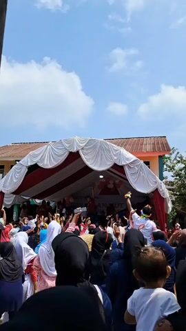 A crowd of people are gathered outside in front of a stage covered by a tent. The tent is draped in red and white fabric. Many of the people are raising their hands. Some are holding up their phones.  The sky is blue with white clouds. There is a building in the background.