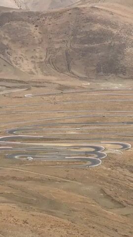 The video shows a long, winding road on a barren, mountainous landscape. The road consists of numerous hairpin turns.  Several vehicles are visible on the road, including what appears to be a white van or SUV.  The terrain is predominantly brown and arid, with some variations in color and texture.  There are some visible utility poles off in the distance. The weather appears to be clear.