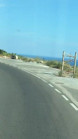The video shows a view from a moving vehicle on a road. The road curves to the left. On the right side of the road, there is a white panel van parked. Further down the road on the right, there is a billboard with an advertisement. Beyond the billboard, there are buildings and a hillside with trees. In the distance, the sea is visible. The sky is clear and blue. The video captures the passing scenery of a coastal road.