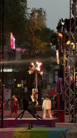 This is a video of an outdoor performance. There are three people on a stage. The person in the middle is an acrobat who is hanging upside down from a pole and swinging. The person on the left is wearing a red costume and is standing still. The person on the right is wearing a white costume and is also standing still. In the background, there are buildings, trees, and lights. The sky is dark, indicating that it is either evening or night. There are also some metal structures that appear to be part of the stage setup or lighting rig.