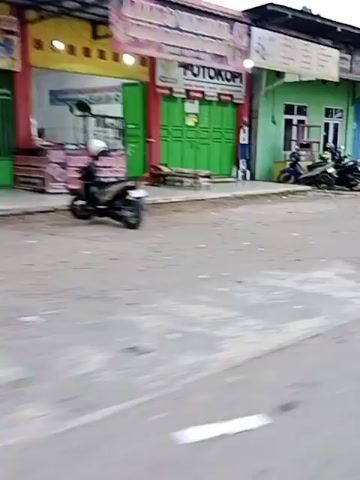 The video shows a street view, likely from a moving vehicle. In the foreground, a blurry palm tree trunk is visible. To the left, there are storefronts. One has green metal shutters and a sign that reads "FOTO KOPI." A person is partially visible next to it. Further down the street, there is a yellow kiosk with a person standing near it, holding a bag. Another kiosk, blue in color, is visible further away. A box of some kind is on the sidewalk near the blue kiosk. In the background, there are buildings, vegetation, and parked motorcycles. The overall impression is of a busy street scene in an urban or semi-urban area, possibly in Southeast Asia, given the style of the buildings and the presence of motorcycles. The video is shaky and out of focus in many parts, suggesting it was recorded while in motion.