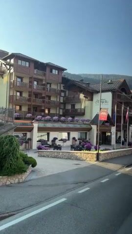 A panning shot captures the exterior of a multi-story hotel with a dark wood and cream facade, balconies adorned with purple flowers, and a paved courtyard with outdoor seating. A street runs in front of the hotel. The sky is clear blue. The camera angle is slightly elevated, suggesting it is mounted on a vehicle. There are flags flying in front of the hotel.