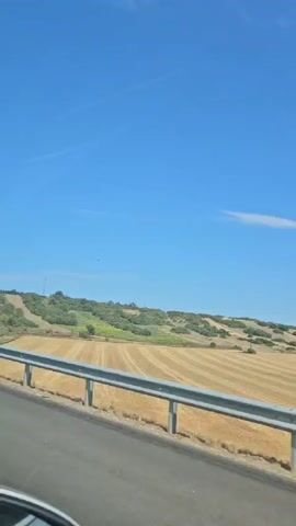 The video shows a view from a moving vehicle, looking out over a rural landscape under a clear blue sky. A highway guardrail runs along the bottom of the frame. Beyond the guardrail, there are fields with dry, golden stubble, suggesting a recently harvested crop. In the middle distance, there are some green trees and bushes, with gently rolling hills in the background. A few scattered white clouds are visible in the sky. The overall impression is of a sunny day in a countryside setting.