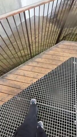 The video begins with a close-up, first-person view of someone's feet standing on a metal grate, with a wooden walkway and metal railing in the foreground. The railing and walkway appear damp, likely from mist. The camera then pans up and to the right, revealing a vast, powerful waterfall plummeting into a canyon.

The waterfall dominates the center of the frame, with white, churning water. The surrounding landscape is rugged and desolate, characterized by brown and green mossy ground, with patches of snow visible in the distance. The sky is heavily overcast with dark grey clouds. The air is thick with mist from the waterfall, creating a hazy atmosphere, particularly noticeable near the water's edge and on the railing. The scene captures the raw power and scale of a natural wonder, likely Dettifoss in Iceland, given the location context. There is no visible text.