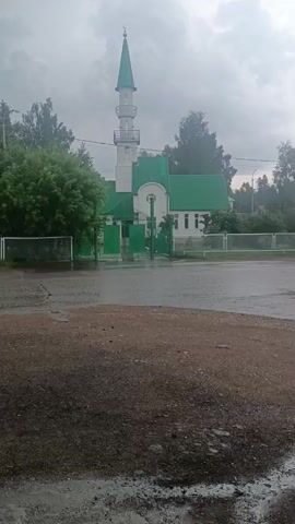 The video shows a mosque with a white minaret topped with a green spire. The mosque itself has a green roof and white walls. A green gate stands in front of the mosque, and a fence surrounds the property. The ground in front of the mosque appears to be wet, possibly from rain. Trees are visible on either side of the mosque. The sky is overcast.
