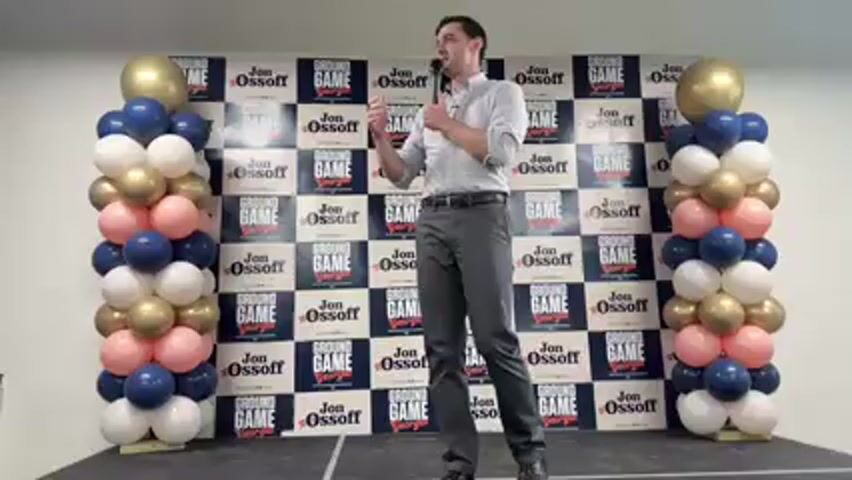 A man stands on a stage or raised platform, speaking into a microphone. He is wearing a light-colored button-down shirt and dark pants, holding the microphone in his left hand and gesturing with his right. Behind him is a checkered backdrop primarily blue and white, featuring the text "JON OSSOFF" and "GROUND GAME GEORGIA" repeated multiple times. "ELECTION.COM" is also visible in smaller text. Two tall columns of balloons flank the backdrop, with a color scheme of gold, dark blue, white, and a pale pink or rose gold. The balloons are arranged in alternating blocks of color. The setting appears to be an indoor political campaign event or rally.