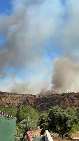A plume of smoke billows into a blue sky with scattered clouds. The smoke appears to originate from a hillside covered in sparse vegetation and rocky outcrops. A structure, possibly a building, is visible on the crest of the hill amidst the smoke. In the foreground, a man wearing a red cap and an orange shirt stands near some greenery, partially obscured by a green tarp. A white railing is also visible in the foreground. The overall scene suggests a wildfire in progress on a hill overlooking a more vegetated area.